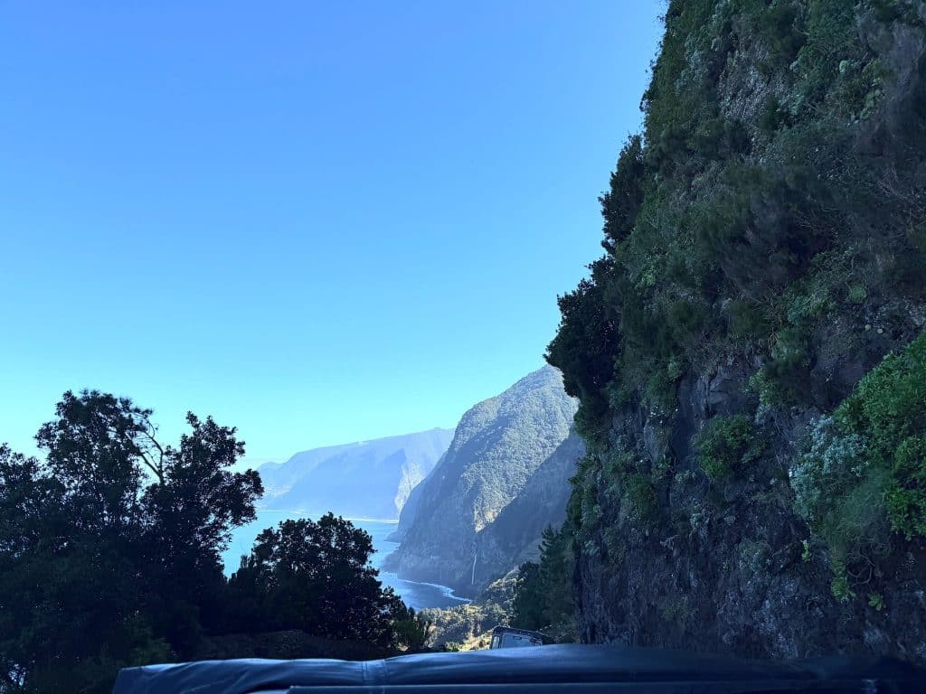 Eine bewaldete Felswand über der Straße, in der Ferne die Klippen der Nordküste und der Ozean, am unteren Bildrand das Dach eines Jeeps