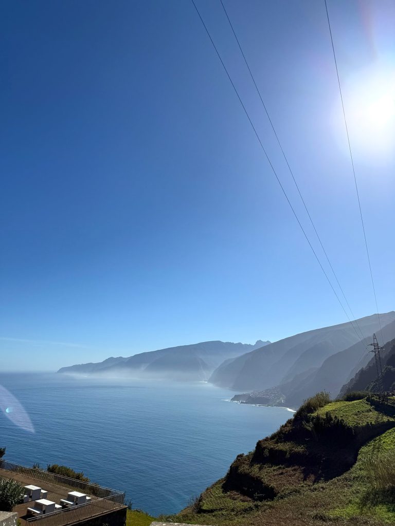 Madeiras Nordküste von oben: steile Hänge, die zum Ozean abfallen, ein Ort auf einer flachen Landzunge, Dunst und Sonne, rechts Stromleitungen