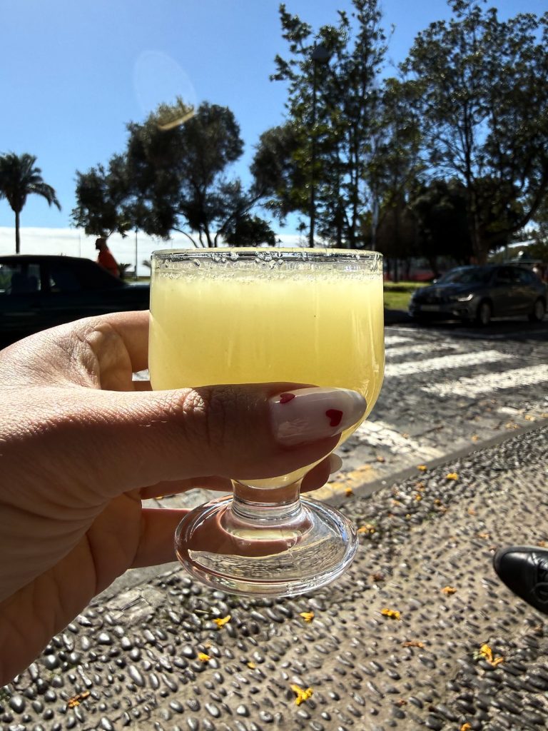 Eine Hand hält ein Stielglas mit hellgelbem Poncha, dahinter eine Straße mit Palmen