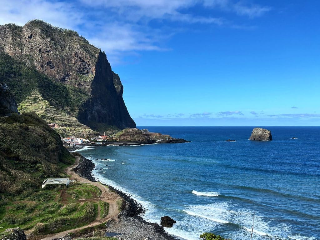 Ein Kieselstrand und Wellen an der Küste von Porto da Cruz, dahinter der Penha d’Águia