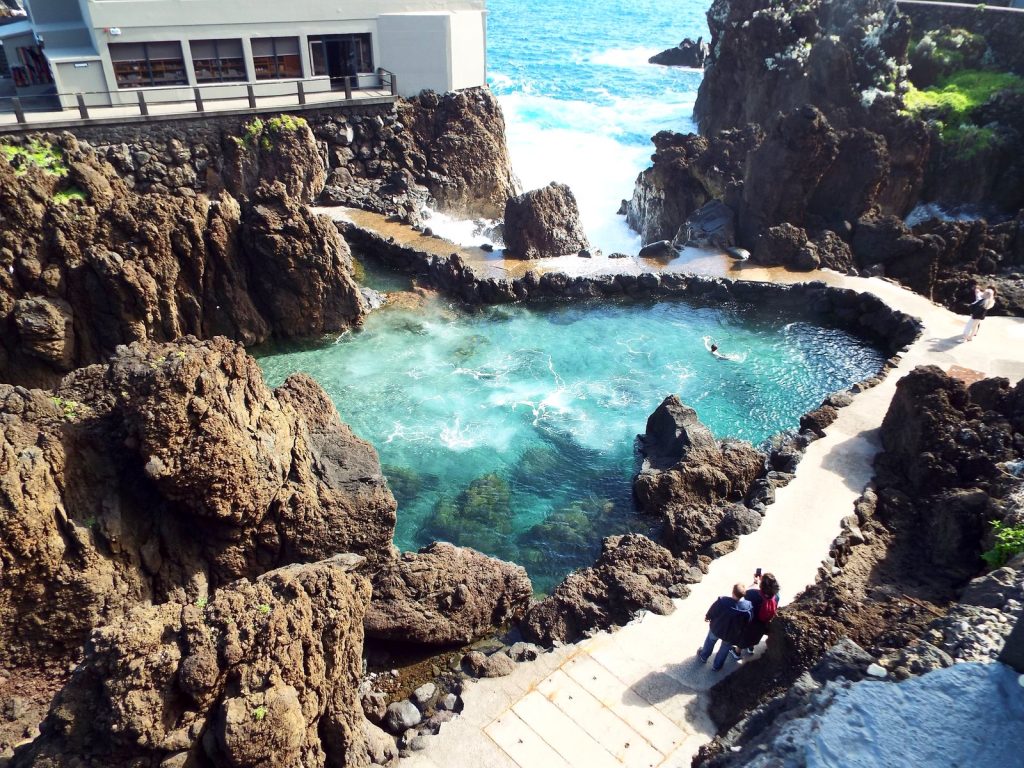 Ein Naturpool mit türkisfarbenem Wasser zwischen schwarzen Vulkanfelsen, am Ufer ein Betonweg und ein Gebäude, hinter den Felsen die Wellen des Ozeans