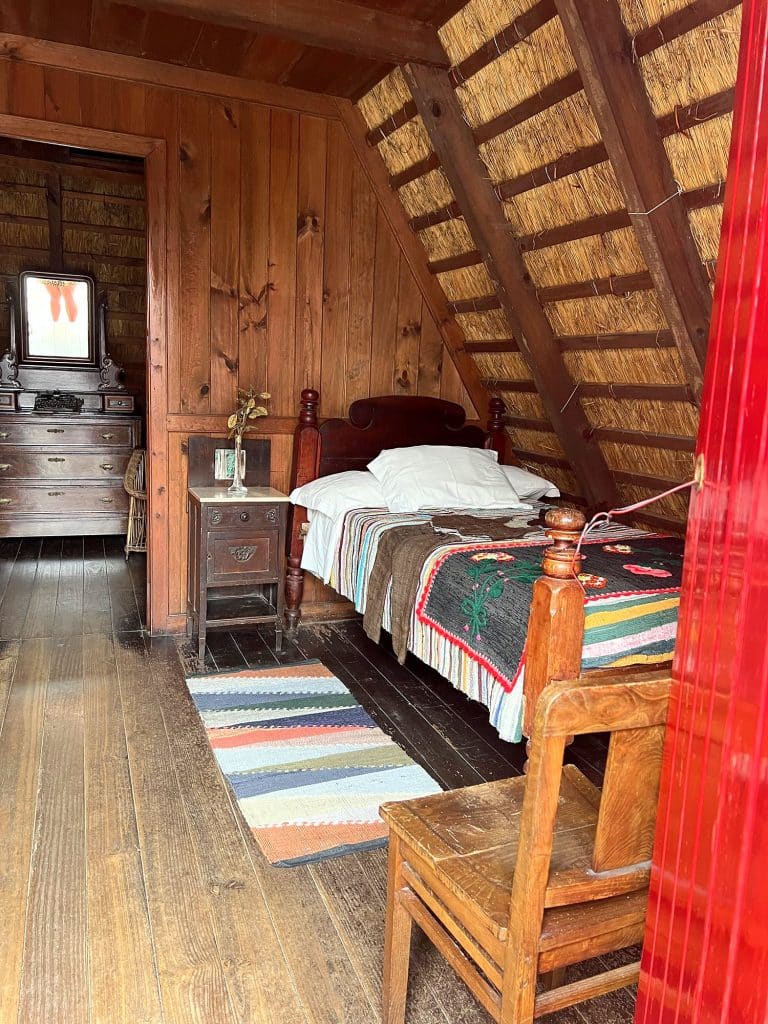 Im Inneren eines Strohdachhauses: Holzbett mit bunter Tagesdecke, gestreifter Teppich, Stuhl und Kommode mit Spiegel