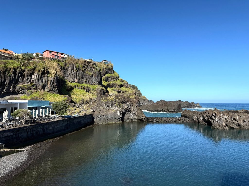 Ein ruhiger Naturpool unter einer Felsklippe mit Häusern obendrauf, am Pool ein Pavillon mit Sonnenschirmen, im Hintergrund der Ozean