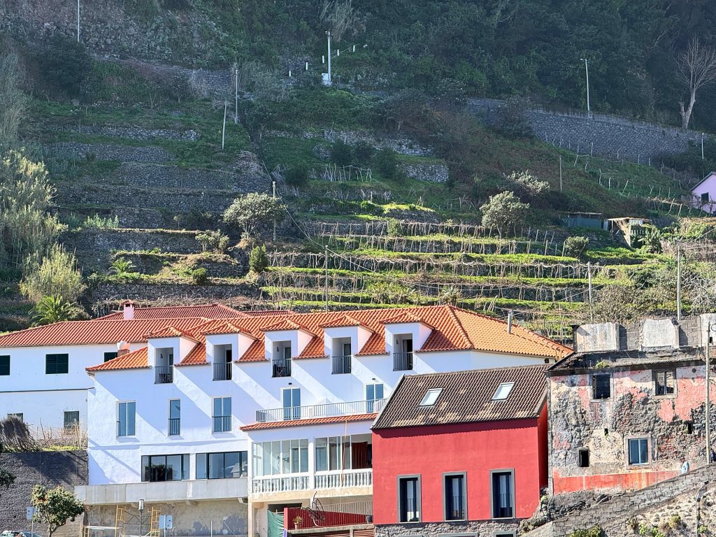 Ein weißes Gebäude mit orangefarbenem Dach, ein rotes Haus und ein altes Gebäude mit abblätternden Wänden unter Terrassen am Hang