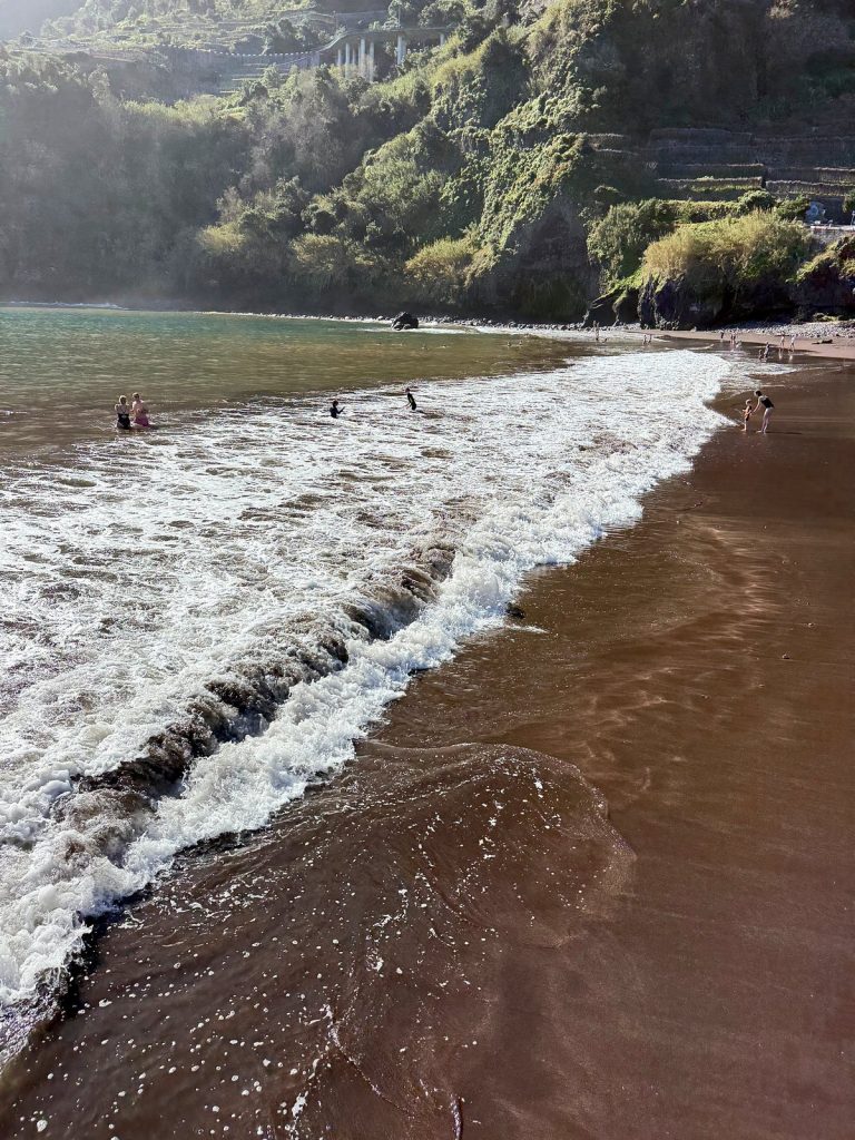 Eine schäumende Welle läuft auf den dunklen Sand, dahinter grüne Klippen in leichtem Dunst und einige Menschen im Wasser