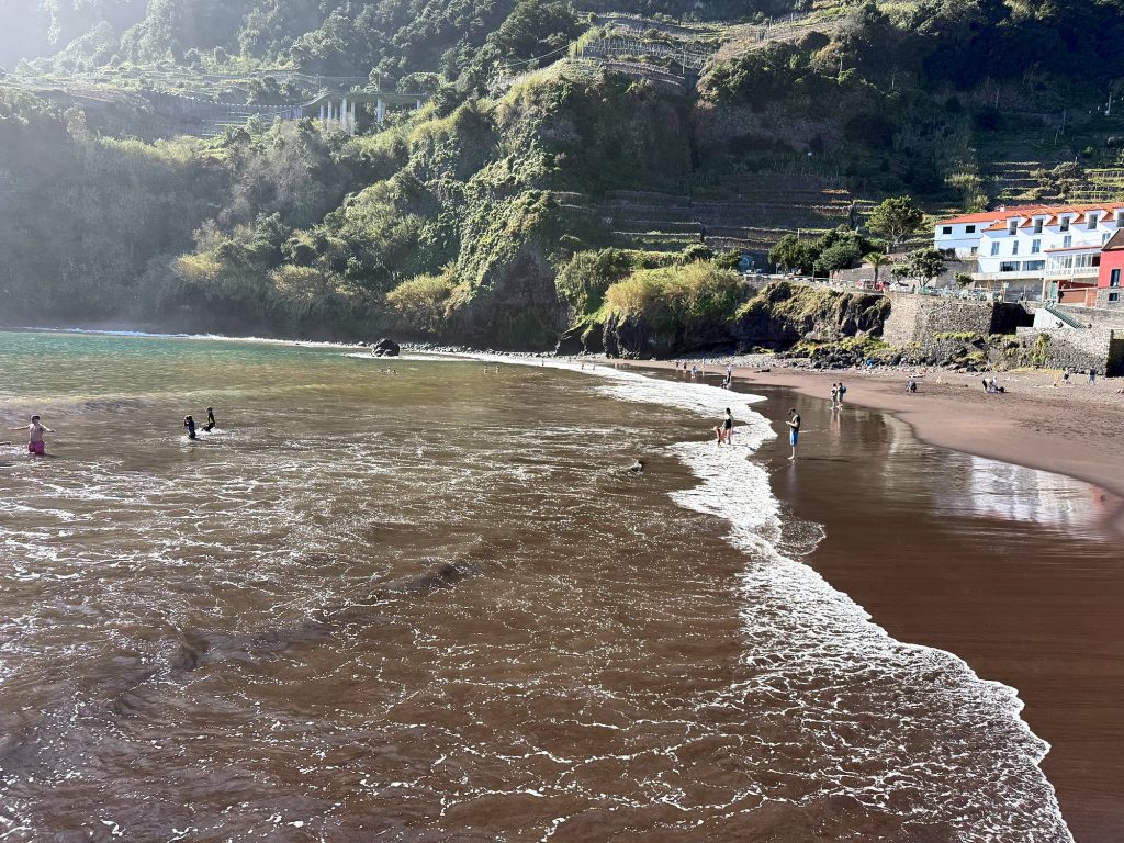 Wellen rollen auf einen Strand mit dunklem Sand, dahinter grüne Hänge und weiße Häuser, im Wasser kleine Figuren von Badenden