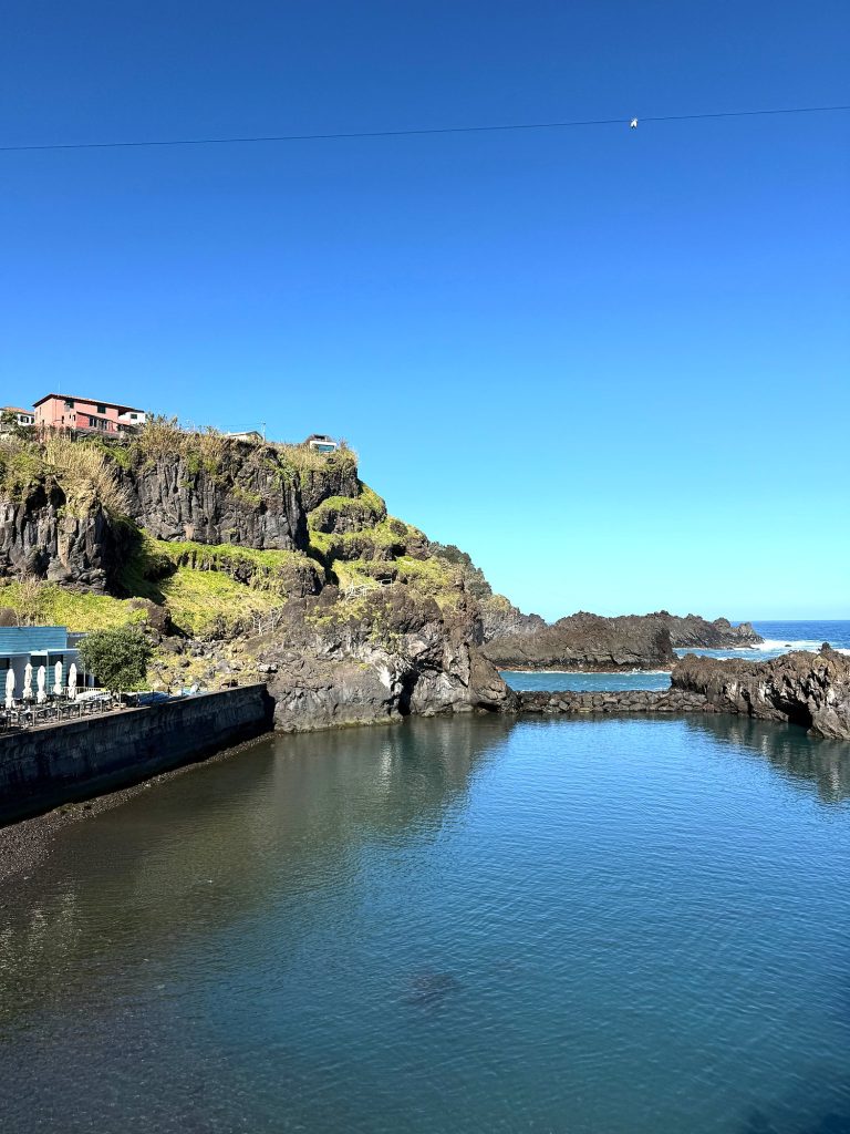 Ein Naturpool mit ruhigem Wasser zwischen Vulkanfelsen, darüber eine Klippe mit einem roten Haus, links Sonnenschirme eines Restaurants