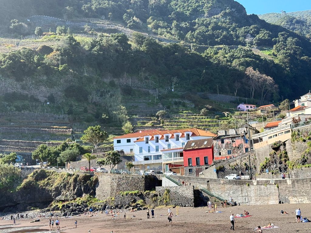 Strand mit dunklem Sand unter einem grünen Terrassenhang, am Ufer ein weißes Gebäude mit orangefarbenem Dach und ein rotes Haus, kleine Figuren am Strand