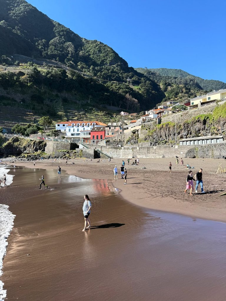 Menschen spazieren über einen Strand mit dunklem Sand, dahinter ein weißes und ein rotes Haus, Hafenmauern und grüne Terrassenhänge