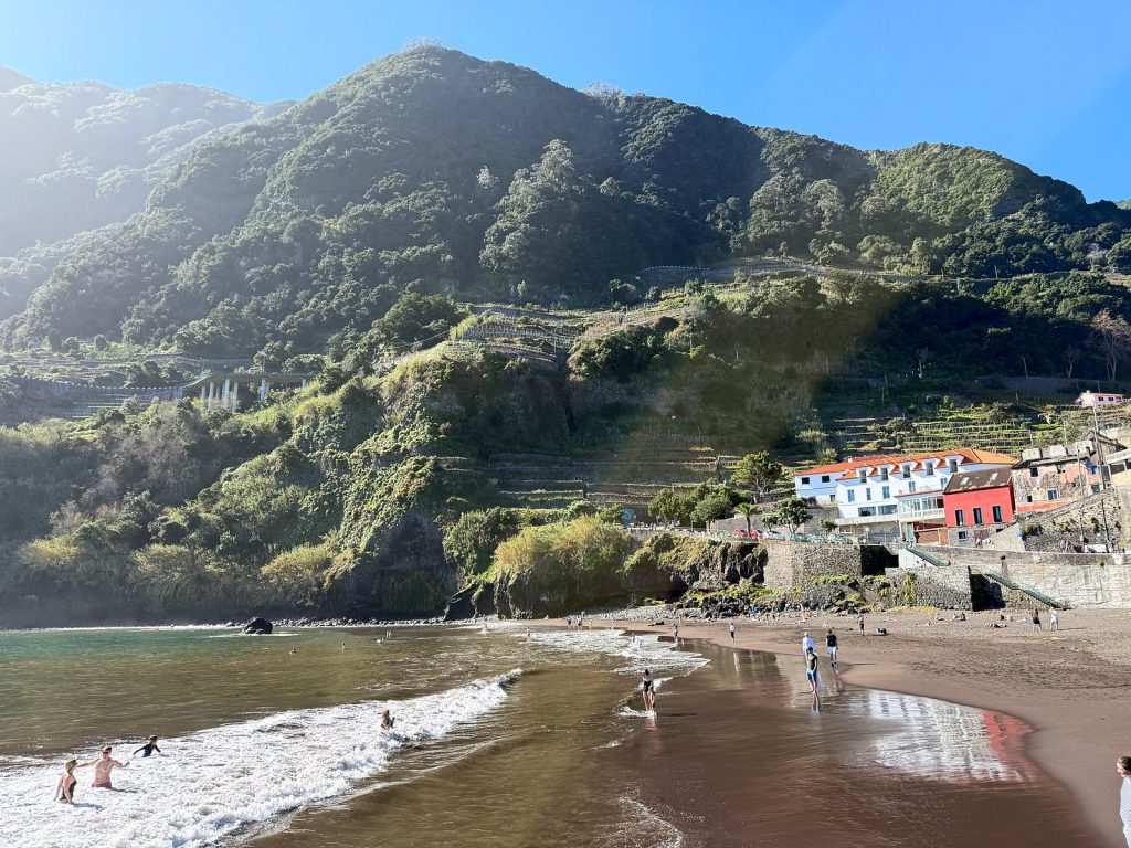Ein Strand mit dunklem Sand unter einem steilen, grünen Terrassenberg, Menschen baden in den schäumenden Wellen am Ufer, rechts ein weißes und ein rotes Haus