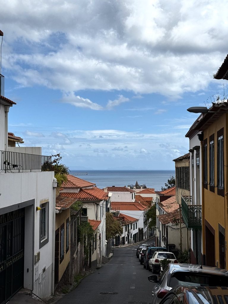 Eine steile Gasse mit geparkten Autos, die zum Meer hinunterführt, rechts ein gelbes Stadthaus