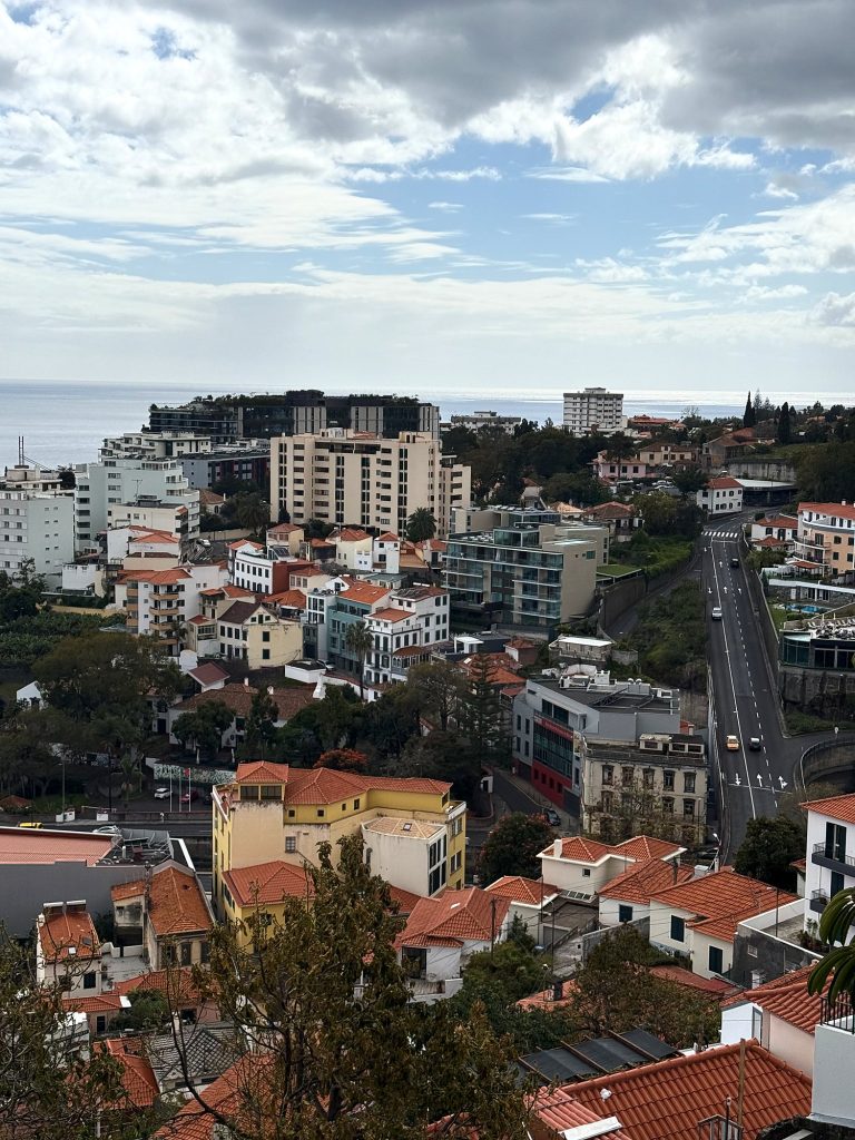 Blick hinunter auf den Westen von Funchal: Stadthäuser mit orangefarbenen Dächern, eine bergab führende Straße, hohe Hotels und das Meer unter bewölktem Himmel